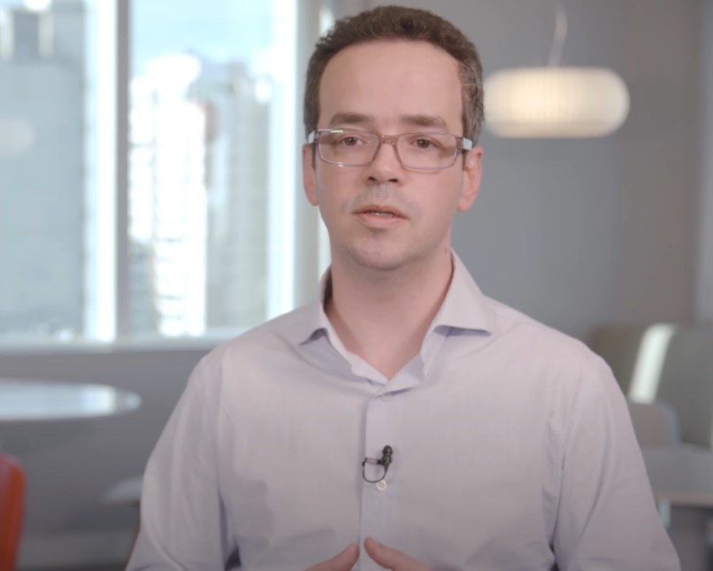 Dr. Caio Nunes, médico usando camisa social clara e óculos, falando em um ambiente profissional com fundo desfocado.