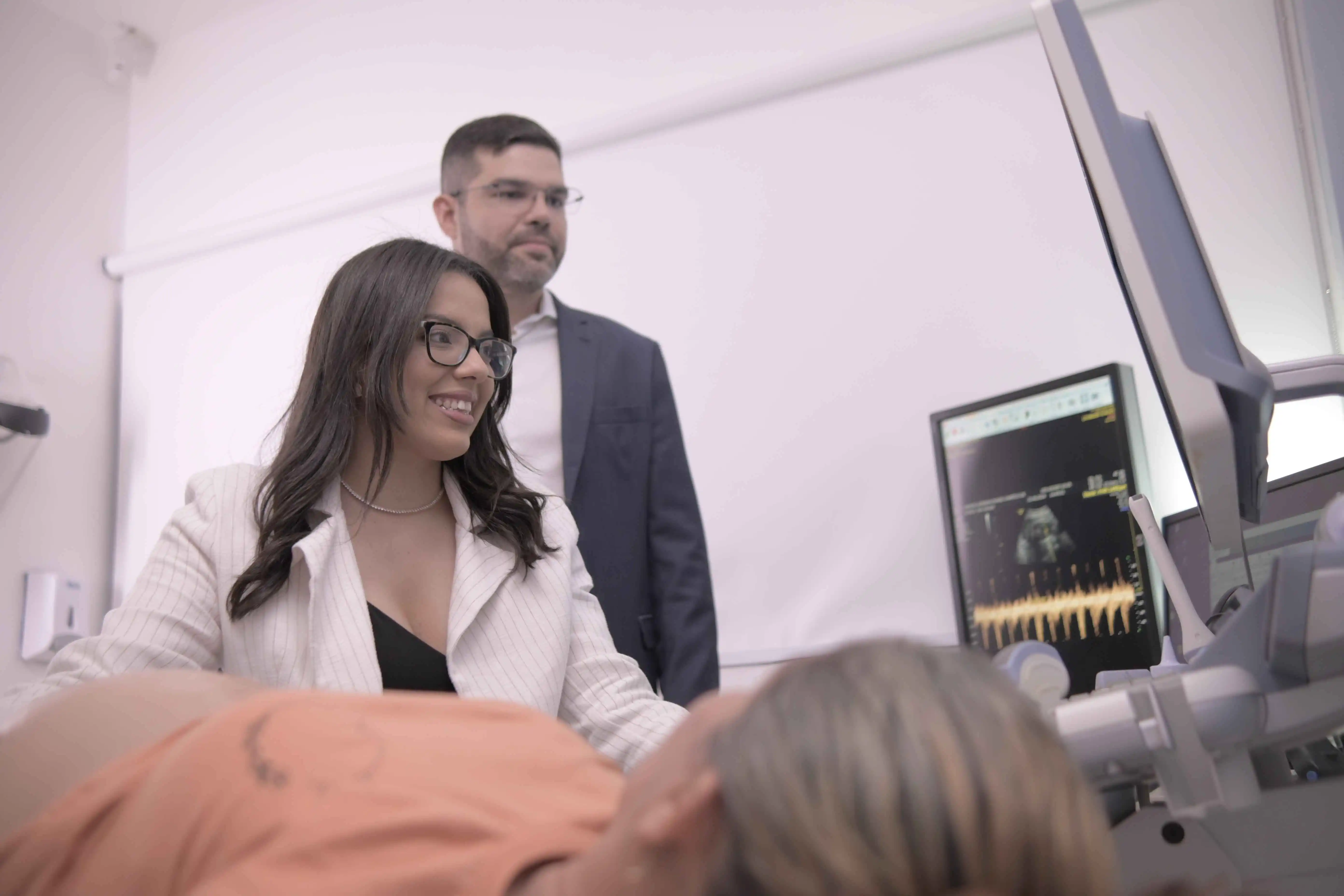 Médica realizando exame de ultrassonografia obstétrica em paciente, com médico observando ao fundo.