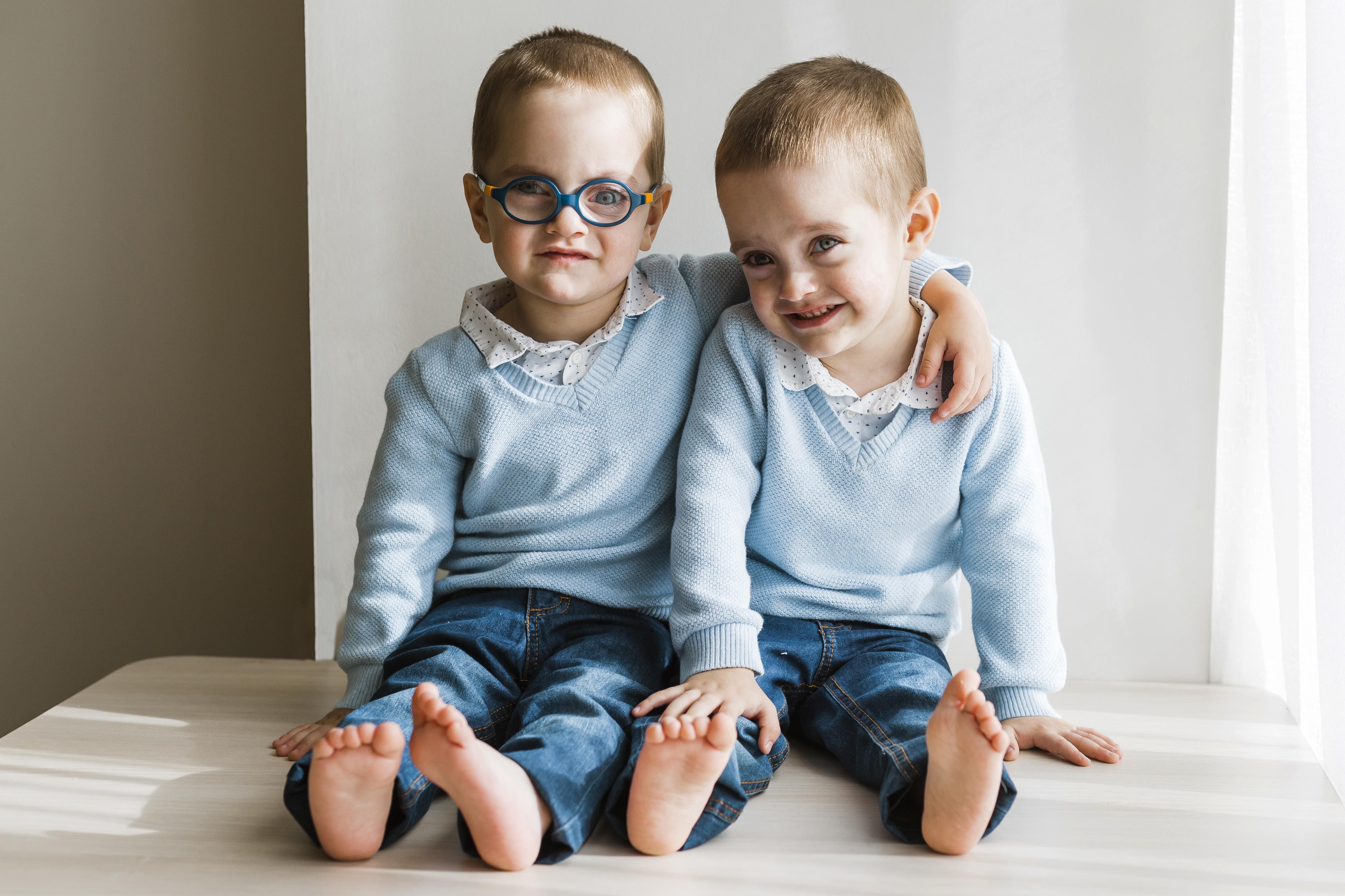 Dois meninos gêmeos com roupas idênticas — suéter azul claro, camisa branca e jeans — sentados juntos, um deles usando óculos azuis e o outro sorrindo com o braço ao redor do irmão.